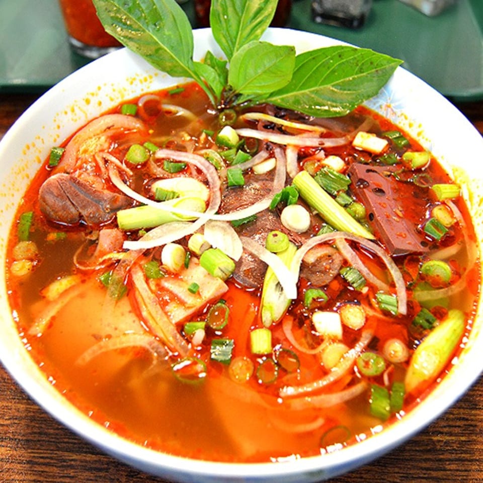 Close-up of a vibrant red Vietnamese soup with meat, noodles, onions, and green herbs.