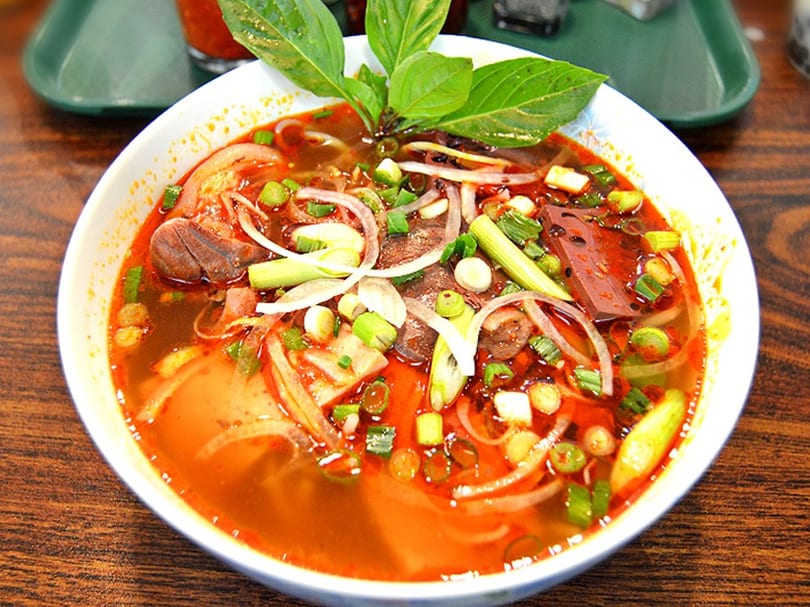 Close-up of a vibrant red Vietnamese soup with meat, noodles, onions, and green herbs.