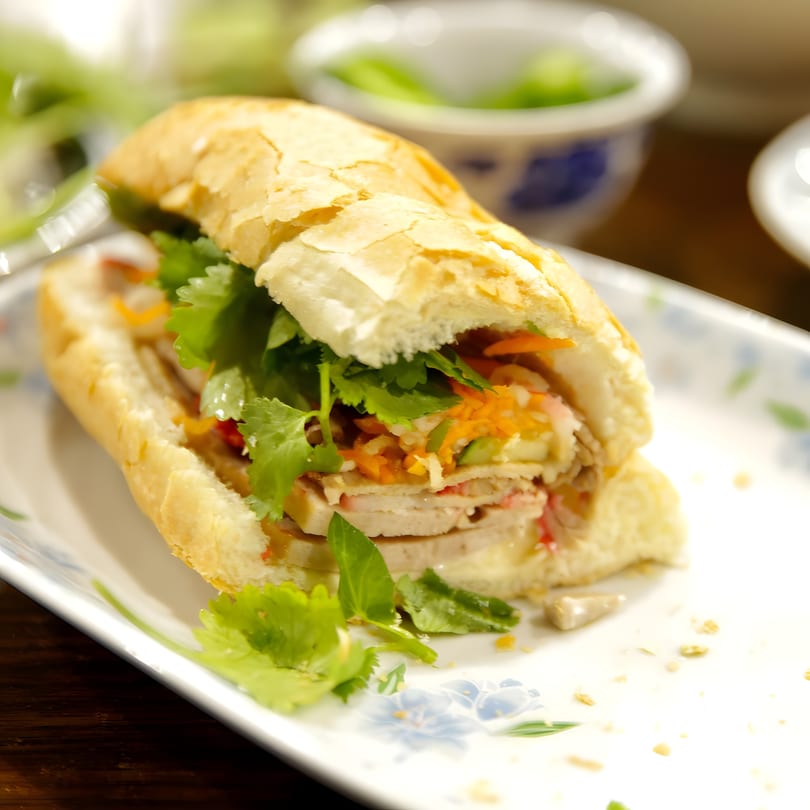 A Vietnamese bánh mì sandwich, filled with meat, pickled vegetables, and fresh herbs, on a white plate.