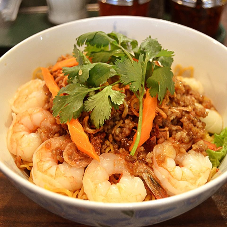 A white bowl filled with shrimp, noodles, minced meat, carrots, and cilantro on a wooden table.