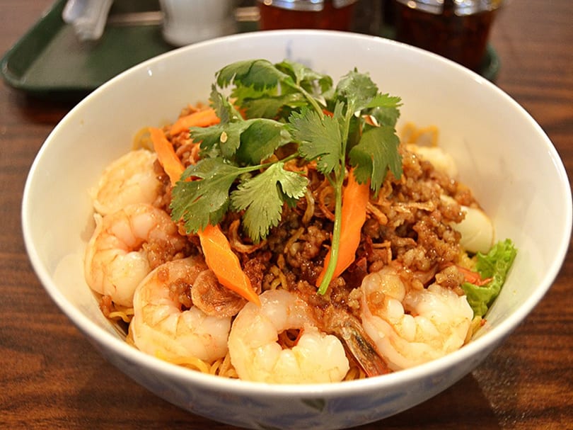 A white bowl filled with shrimp, noodles, minced meat, carrots, and cilantro on a wooden table.