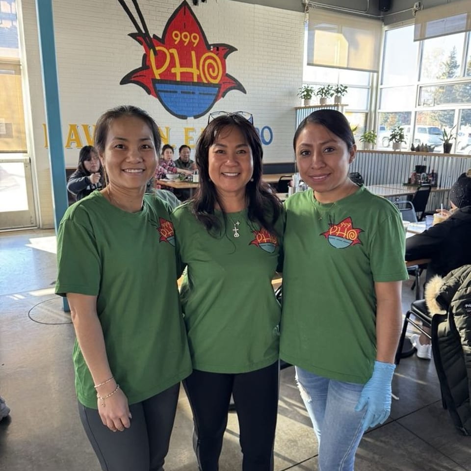 Three smiling women in green Pho 999 shirts stand in a brightly lit restaurant dining area.