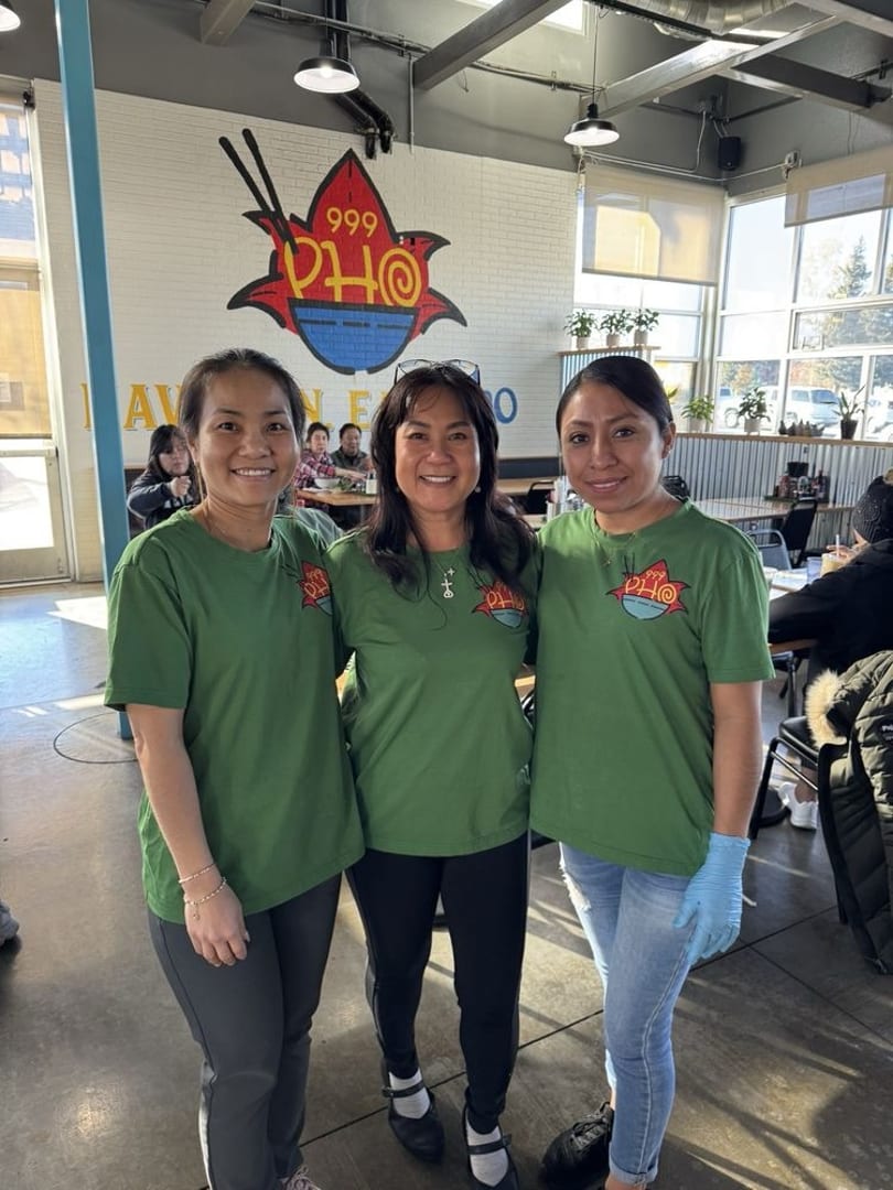 Three smiling women in green Pho 999 shirts stand in a brightly lit restaurant dining area.